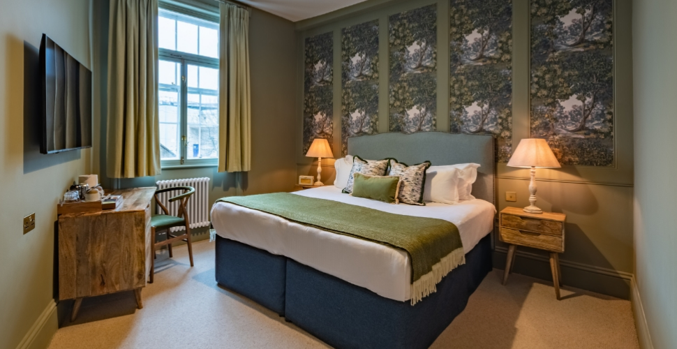 Cosy bedroom with a double bed, white linens, an olive-green throw and patterned cushions. Woodland-patterned panels decorate the wall behind the upholstered headboard, with warm lamps on either side. A wooden desk and chair sit beneath a curtained window, opposite a wall-mounted television. Cosy bedroom with a double bed, white linens, an olive-green throw and patterned cushions. Woodland-patterned panels decorate the wall behind the upholstered headboard, with warm lamps on either side. A wooden desk and chair sit beneath a curtained window, opposite a wall-mounted television.